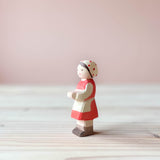 Die Ostheimer Spielfigur Heidi, bemalt mit rot als Kleid und weißer Schürze, außerdem Trägt sie ein rot getupftes Häubchen. Sie steht auf einer Holzplatte und schaut nach links.