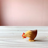Die Ostheimer Huhn Holzfigur zum Spielen ist braun, steht auf einer Holzplatte und hat den Kopf geneigt, als würde es Futter picken. Das Huhn hat einen roten kleinen Kamm.