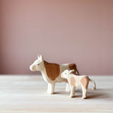 Ostheimer Spielfiguren aus Holz. Die Kuh mit braunen Flecken steht mit erhobenem Kopf und Blick nach links. Ein kleines Kalb steht seitlich mit Blick zur Kuh daneben.