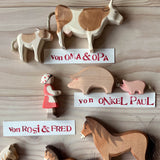 Ostheimer Spielfiguren aus Holz: Kalb, Kuh, Figur Tochter, Schwein, Ferkel und Pferd braun. Daneben liegen Namensschilder, die erklären, wer die Figuren geschenkt hat, zum Beispiel "von Oma und Opa".