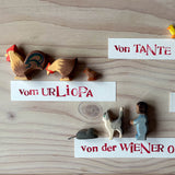 Ostheimer Spielfiguren aus Holz: Hahn, Huhn, dunkles Küken, gelbes Küken, Maus, Katze und ein Kind Spielfigur. Daneben liegen Namensschilder, die erklären, wer die Figuren geschenkt hat, zum Beispiel "vom Urliopa".