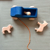 Ein Bild von oben auf ein blaues Nachzieh-Holzauto mit Schnur. Auf der Ladefläche liegt eine runde Holzfigur. Auf der karierten Matte liegen ein kleines Naturholzpferd und ein Naturholzhund ohne Bemalung.