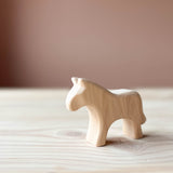 Ein Holzpferd aus hellem Holz ohne Bemaung steht auf einer Holzplatte und ist seitlich nach vorne gerichtet.