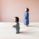 Die Ostheimer Spielfigur Wickelkind steht auf einer Holzplatte und schaut nach links. Im Hintergrund rechts ist unscharf die Ostheimer Holzfigur Mutter zu sehen.