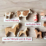 Ostheimer Spielfiguren aus Holz: Kalb, Kuh, Figur Tochter, Schwein, Fohlen braun, Haflinger und Haflingerfohlen. Daneben liegen Namensschilder, die erklären, wer die Figuren geschenkt hat, zum Beispiel "von Oma und Opa".