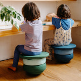 Zwei Kinder sind von hinten zu sehen, sie sitzen auf gestapelten Stapelsteinen, mit je zwei Original Stapelsteinen und einem Balance Board.