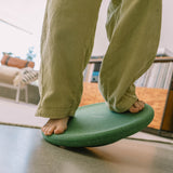 Jemand balanciert auf dem Stapelstein Balance Board in Dunkelgrün. Es sind nur die nackten Füße und eine lange Cordhose bis unterhalb des Knies zu sehen.