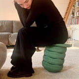 Ein junge Frau, schwarz gekleidet, sitzt auf einem gestapeltem Turm aus einem Stapelstein Balance Board in dunkelgrün ganz oben und vier weiteren dunkelgrünen Stapelstein Original Steinen.