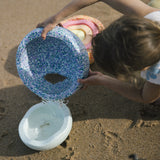 Ein Kind am Strand schüttet Wasser vom Stapelstein Confetti in einen Stapelstein Mini in hellblau.