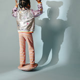 Ein Mädchen balanciert auf einem Stapelstein Balance Board Confetti. Sie wird von hinten beleuchtet und ihr Schatten fällt an die Wand. Im Schatten ist zu sehen, dass sie auch einen Stapelstein auf dem Kopf balanciert.