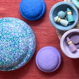 Der Stapelstein® Confetti Single Super Cool liegt neben vier lilafarbenen und blauen Stapelsteinen Mini, zwei sind zu Schalen umgedreht und beherbergen farblich passendes Grapat Spielmaterial.