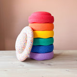 Stapelstein Original Rainbow 6er Set gestapelt auf einem Turm, ganz unten liegt der violette Stein, darüber der blaue, dann der grüne, gefolgt vom gelben, dann orange und ganz oben der rote Stapelstein. Links am Stapelstein Turm angelehnt, ist ein Stapelstein Balance Board Confetti zu sehen, ganz bunt gesprenkelt.