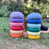 Zwei Türme mit je sechs Stapelsteinen Mini in verschiedenen Farben stehen in der Wiese, dahinter versteckt sich ein Kind liegend und hält den Daumen hoch zwischen den zwei Türmen.