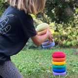 Ein Kind hält einen farblich sortierten Turm aus sechs Stapelsteinen Mini in Pastell, im Begriff diesen auf einen Turm in Regenbogenfarben zu setzen.