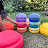 Zwei bunte Stapbel von jeweils sechs Stapelsteinen Mini stehen in der Mitte, ringsherum Stapelsteine Original in der Wiese verteilt.