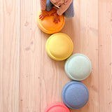 Pastell Stapelsteine sind von oben fotografiert, sie liegen wie ein gebogener Weg auf einem hellen Holzboden. Ganz unten ist ein rosa Stapelstein, gefolgt von einem hellblauen, dann ein mintgrüner, dann blassgelb und dann pastellorange. Auf dem orangen Stapelstein stützt sich ein Kind ab.