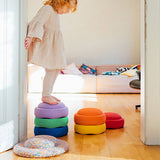 Ein Stapelstein Bundle Classic in Regenbogenfarben in drei verschiedenen Teilen, mal drei, mal zwei gestapelt, mal ein loser Stein, daneben liegt ein Confetti Board. Ein Kind balanciert darüber.