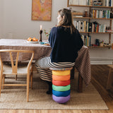 Eine Frau sitzt auf einem Hocker, der aus einem Stapelstein Regenbogen Set Classic und einem Confetti Board zusammengebaut ist.