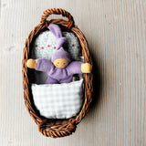 Das Wichtelnest von Kokomoo in lichtblau: ein Weidenkörbchen als Puppenbett, in dem ein lila Nanchen Wichtel in einer hellblau gemusterten Bettwäsche kuschelt.