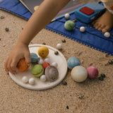 Die Planeten, Monde und Sterne von Grapat Dear Universe liegen auf einem Holzteller am Strand, ein Kind greift gerade nach Jupiter.