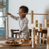 Ein Kind spielt mit Grapat Woodland Bausteinen und legt gerade eine Holzleiste auf den Bausteine-Turm. Davor am Tisch stehen Teller mit Kuchen.