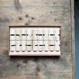 18 Holzzylinder, jeder zart bemalt, liegen sortiert in einem Karton. Man kann die Schlitze in de Zylindern erkennen.