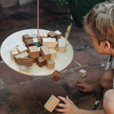 Grapat "The Pendulum" - eine an einem Stab baumelnde Holzplatte mit verschieden platzierten Quader darauf, von oben fotografiert, einige Holzbausteine fallen schon runter. Ein Kind hockt daneben.
