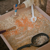 Verschiedene Samen und Körner liegen in einer flachen Holzwanne, ein Kind stampft diese mit einem Werkzeug von Grapat "Tools" Handwerkzeug Spielset aus Holz.
