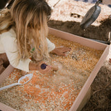 Verschiedene Samen und Körner liegen in einer flachen Holzwanne, ein Kind zerkleinert diese mit einem Werkzeug von Grapat "Tools".