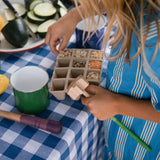 Ein Kind sortiert verschiedene Körner und Samen in eine Anzuchtschale, in der anderen Hand hält es ein Grapat Tool Werkzeug aufs Holz.