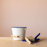 Ein kleiner, weißer Emaillekübel mit blauem Rand, ein kleiner Handbesen und eine kleine, blaue Kehrschaufel aus Metall.