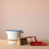 Ein kleiner, weißer Emaillekübel mit blauem Rand, ein kleiner Handbesen aus Holz mit Naturborsten und eine kleine, rote Kehrschaufel aus Metall.