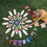 Ein wunderschön aufgelegtes blütenförmiges Mandala in der Wiese. Die blätterförmigen Holzplatten von Grapat liefern die Basis, darauf verteilt ein Kind bunte Mandalasteine. Die verschiedenen Mandalas stehen in ihren Boxen bereit.