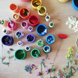 Die bunten Holzbecher von Grapat "Nest Bowls" in den verschiedenen Größen in Schattierungen der Regenbogenfarben. In und um die Becher liegen Blumen.