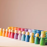 Carlas Nins 120-teiliges Set mit 12 Nins Holzfiguren in Regenbogenfarben stehen nebeneinander, dahinter die Plättchen und Ringe in den passenden Farben.