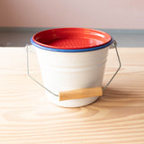 Auf den kleinen weißen Eimer für Kinder liegt ein rotes Sieb aus Metall auf, das ist robustes Sandspielzeug.