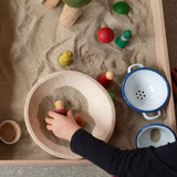 Ein Kind spielt mit der Sandwanne, darin ist eine Holzschale, ein Emaille Nudelsieb, eine kleine Emailleschale und kleine Holzfiguren zum Spielen.