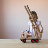 Ein Kind spielt mit dem Fagus Feuerwehr Drehleiterfahrzeug, in der einen Hand die Holzfiguren mit den roten und blauen Köpfen von Fagus.