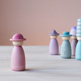 Vorne steht eine Grapat Fancy Nins Spielfigur in rosa mit einem passendem Hut, im Hintergrund weitere Spielfiguren aus Holz in Blau- und Lilatönen.