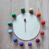 Ein Kreis mit 12 Schälchen aus Holz von Grapat, gefüllt mit 12 Filzbällen von Kokomoo in Regenbogen Farben. In der Mitte liegt eine Lampionblume auf einer kreisförmigen Holzplatte.