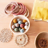 Viele kleine Filzkugeln von Papoose Toys in verschiedenen Farben liegen in einer Weidenholzschale, rundherum liegen gelbe Baumwolltücher, rosa Filzringe und verschiedene Körbe und Untersetzer.
