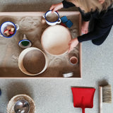 Draufsicht auf ein spielendes Kind, das einen Emaillebecher mit Sand gefüllt in der einen und eine Holzschale in der anderen Hand hält. Das Kind kniet neben einer Sandwanne , die mit Sand, Sieb, Emaille-Trichter und kleinen Gegenständen gefüllt ist. Daneben liegen Besen und Schauferl.