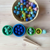 Ein Korb voll mit kleinen Filzkugeln in Blau- und Grüntönen, darunter die fünf Stapelbecher des Grimms Schälchenset "Ozean" nebeneinander aufgestellt und Filzkugeln darin farblich zugeordnet. Ganz unten eine Mini Holzzange zum Greifen.