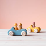 Grimms Holzauto in blau, dahinter das gelbe Holzauto. Im blauen Auto sitzen zwei Spielfiguren aus Holz, im gelben eine.