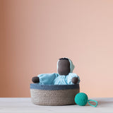 Die blaue Kuschelpuppe "Aqua" von Grimms aus Stoff sitzt in einem Korb mit blauem Rand. Daneben liegt ein türkiser Häkelball mit Schlaufe von Kokomoo.