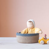 Die gelbe Kuschelpuppe "Sonne" von Grimms aus Stoff sitzt in einem Korb mit blauem Rand. Daneben steht ein kleines, gelbes Holzauto.