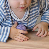 Ein Baby stützt sich auf eine Holzplatte, in der rechten Hand einen lilafarbenen Häkelball von Kokomoo.