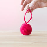 Eine Hand hält den Häkelball mit Schlaufe in der Farbe Kirsche an der Schlaufe hoch.