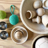 Ein Häkelball mit Schlaufe in Türkis liegt neben Holzschälchen, kleinen Klangschalen und anderen freien Spielmaterialien aus Holz.