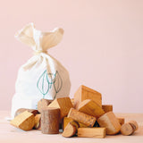 Ein Hügel naturbelassener Bausteine bzw Bauklötze aus Holz - samt Rinde - in ganz unterschiedlichen Formen, liegen auf einer Holzunterlage. Daneben steht ein Stoffbeutel, in dem die Basusteine verpackt sind.